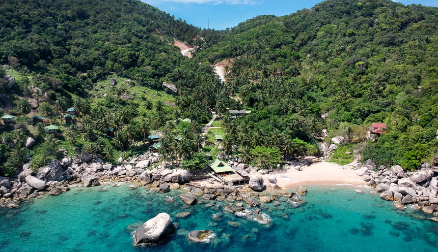 In primo piano le acque turchesi bagnano una spiaggia sabbiosa sulla destra e un gruppo di scogli sulla sinistra. Dietro vegetazione tropicale.