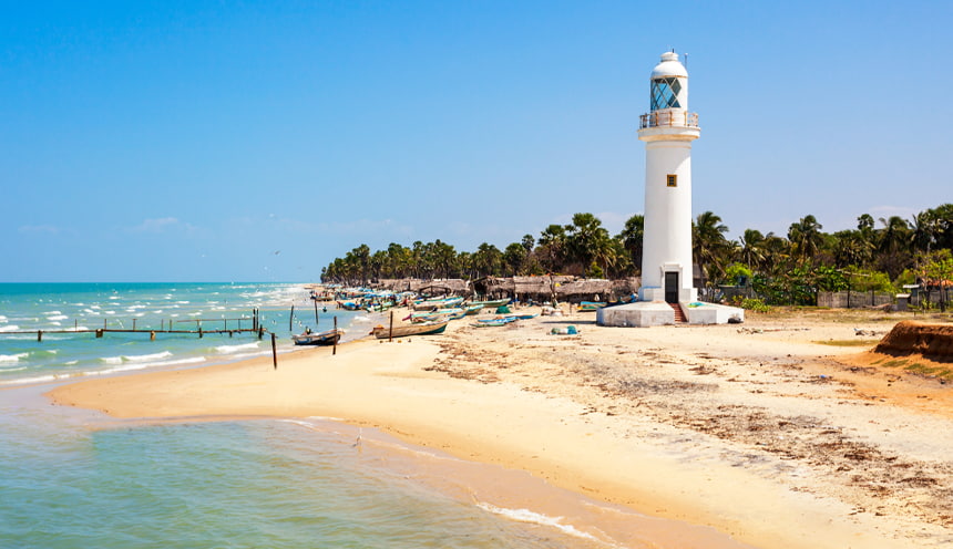 Foto di una spiaggia sulla destra di color giallino e un alto faro bianco. Alle spalle alberi e palme.