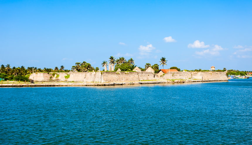 Al centro le mura di un forte coloniale con alcune palme alle spalle. In primo piano il mare azzurro.