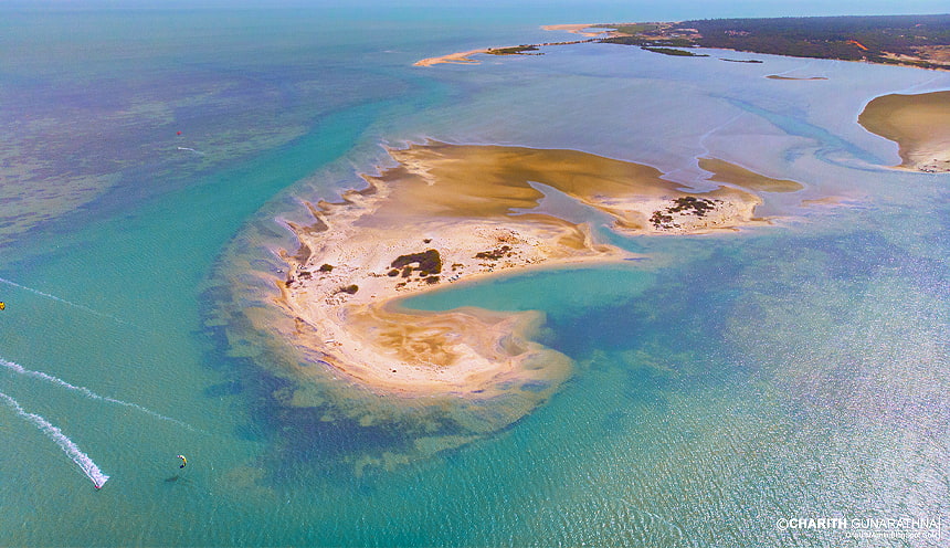 Foto aerea di una piccola isola sabbiosa color giallo-ocra, circondata da acque smeraldine e da due kite-surf