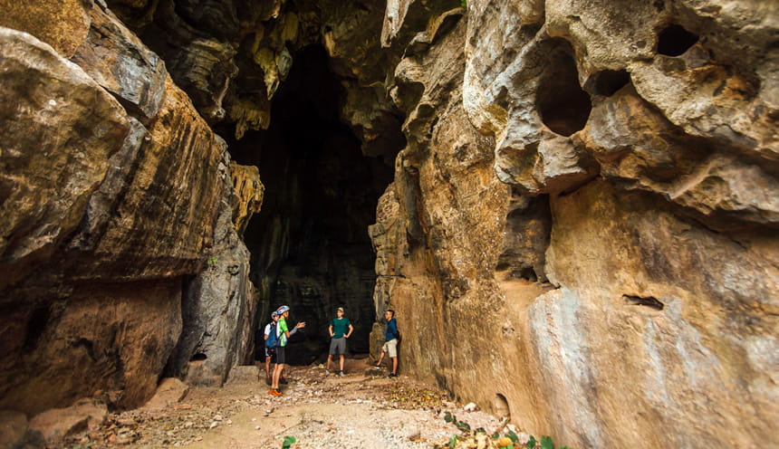 L'ingresso di una caverna dalla alte pareti con al centro tre escursionisti e una guida.
