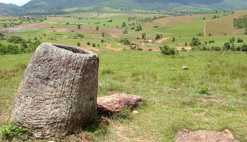 Sulla sinistra una giara in pietra in primo piano si affaccia su una vallata verde con qualche albero e campo coltivato.